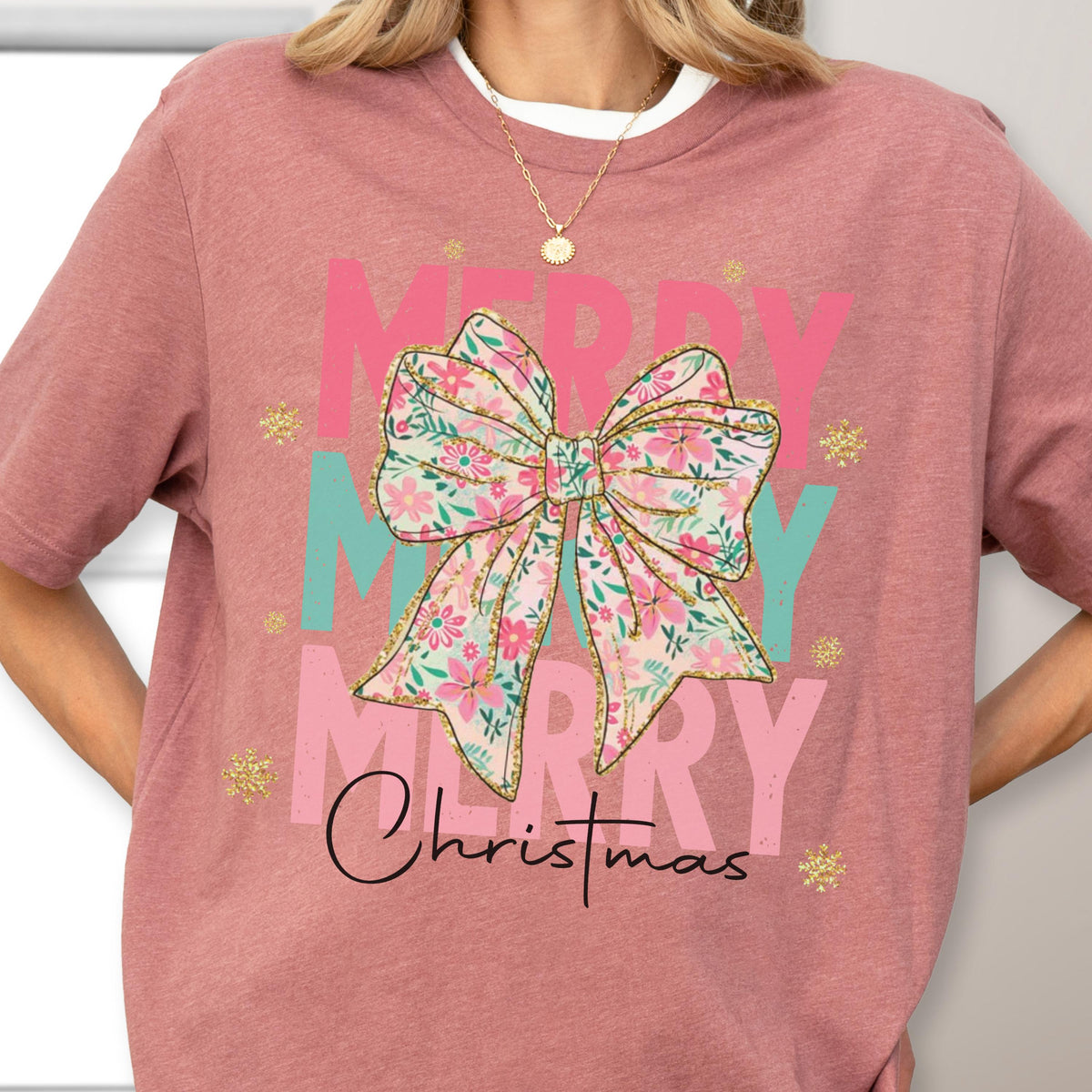 May include: A mauve-colored t-shirt featuring a floral bow design with the words "MERRY" in pink and teal, and "Christmas" in black script. Gold glitter accents and a white crew neck are also visible.