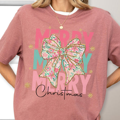 May include: A mauve-colored t-shirt featuring a floral bow design with the words "MERRY" in pink and teal, and "Christmas" in black script. Gold glitter accents and a white crew neck are also visible.