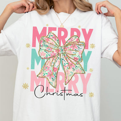 May include: White t-shirt with a festive design. The shirt features the words "MERRY" in pink and teal, with a floral bow graphic in the center. The word "Christmas" is written in black script below the bow, with gold glitter accents.