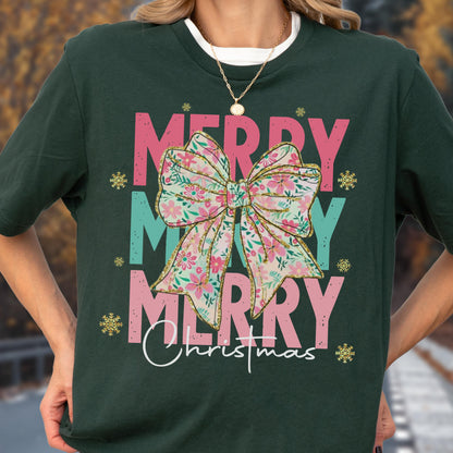 May include: Dark green t-shirt with a floral bow graphic. The words "MERRY MERRY MERRY Christmas" are printed in pink, teal, and white. Gold snowflakes accent the design. A festive holiday tee.