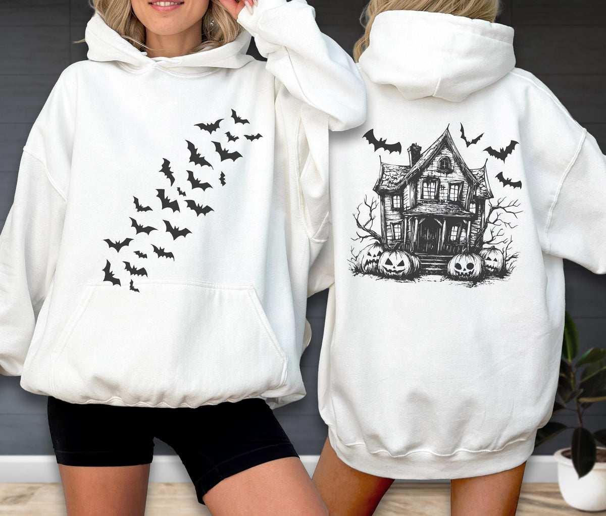 May include: A white hooded sweatshirt featuring a black graphic of a swarm of bats on the front. The back displays a detailed black illustration of a haunted house surrounded by bare trees and several carved jack-o'-lantern pumpkins.
