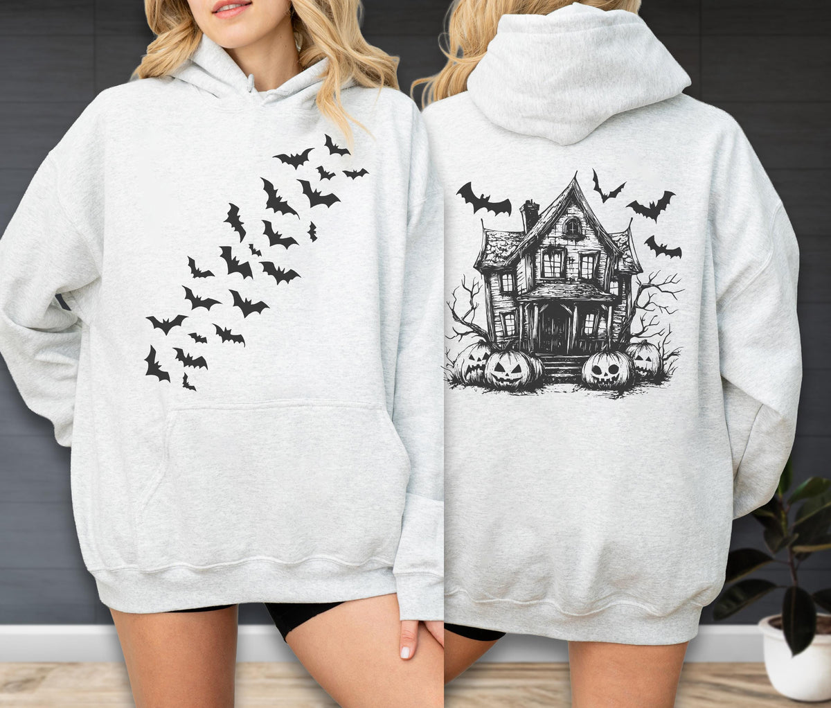 May include: A light gray hooded sweatshirt featuring a black graphic of a swarm of bats on the front. The back of the sweatshirt displays a black ink-style illustration of a haunted house surrounded by bare trees and several carved jack-o'-lanterns.