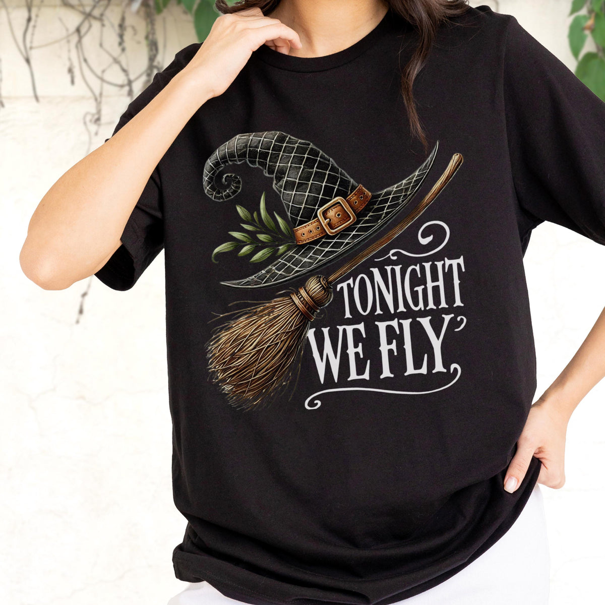 May include: A black t-shirt featuring a graphic print of a black witch hat with a brown leather buckle and green leaves, paired with a wooden broomstick. The text "TONIGHT WE FLY" is printed in white lettering below the hat and broomstick design.