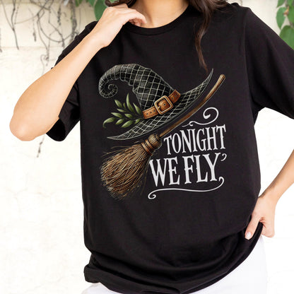 May include: A black t-shirt featuring a graphic print of a black witch hat with a brown leather buckle and green leaves, paired with a wooden broomstick. The text "TONIGHT WE FLY" is printed in white lettering below the hat and broomstick design.