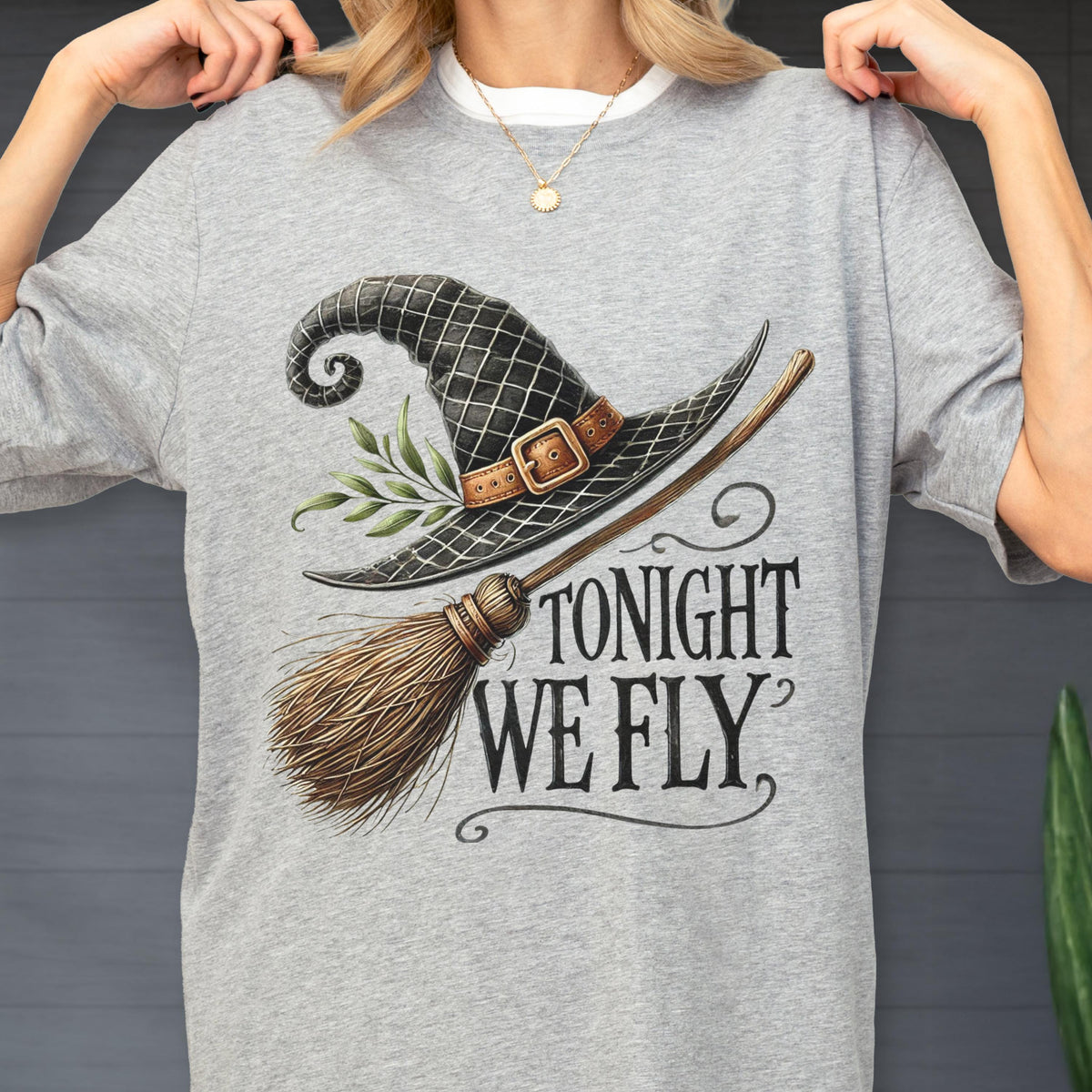 May include: A heather gray t-shirt featuring a graphic of a black witch hat with a brown buckle and green leaves, paired with a wooden broomstick. The text TONIGHT WE FLY is printed in a black serif font below the hat and broom illustration.