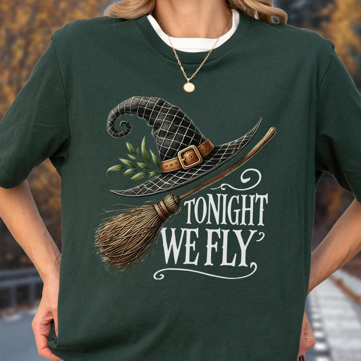 May include: A dark forest green short-sleeved t-shirt featuring a graphic print of a black witch hat with a brown buckle, a small green leafy sprig, and a wooden broomstick. The white text below the graphic reads, "TONIGHT WE FLY."