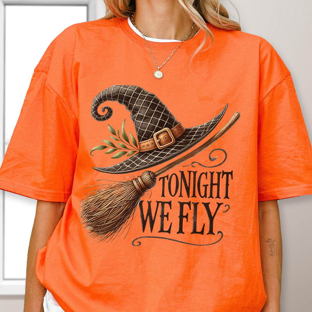 May include: An orange t-shirt featuring a graphic of a black witch hat with a brown buckle and green leaves, paired with a wooden broomstick. The text "TONIGHT WE FLY" is printed in a black, stylized serif font below the witch hat illustration.