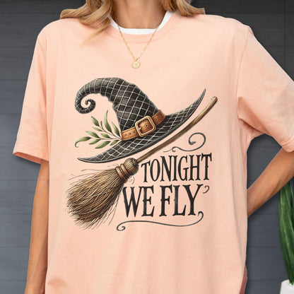 May include: A peach-colored t-shirt featuring a graphic of a black witch hat with a brown buckle and green leaves, paired with a wooden broomstick. The text TONIGHT WE FLY is printed in a black serif font below the hat and broomstick illustration.