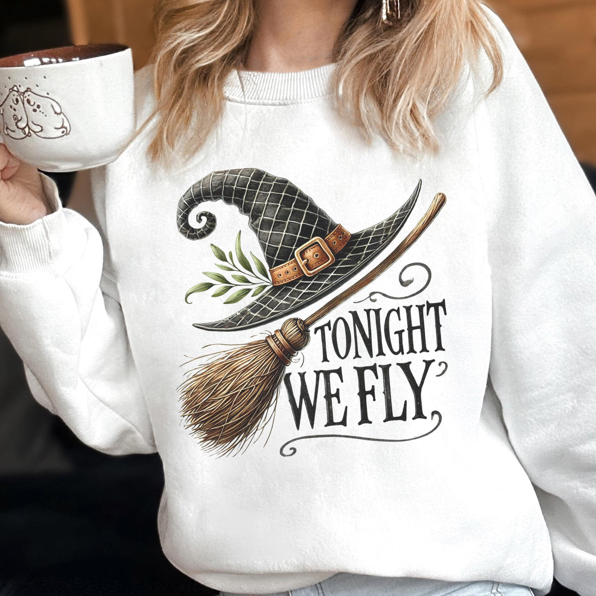 May include: A white crewneck sweatshirt featuring a graphic print of a black witch hat with a brown buckle, green leaves, and a wooden broomstick. The text "TONIGHT WE FLY" is printed in black lettering below the hat and broomstick illustration.