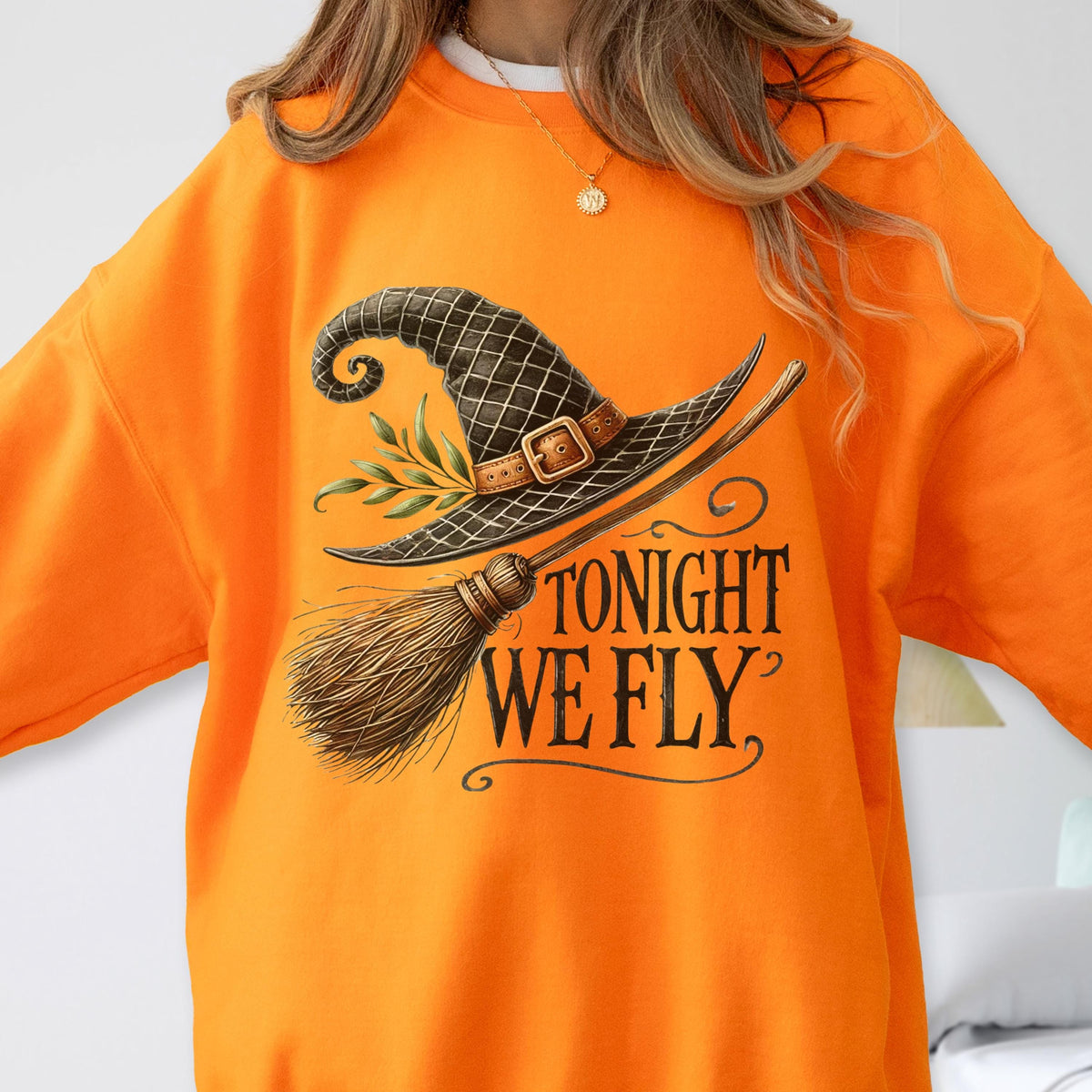 May include: A bright orange crewneck sweatshirt featuring a graphic print of a black witch hat with a brown buckle and green leaves, paired with a wooden broomstick. The text TONIGHT WE FLY is printed in a dark, stylized serif font below the hat.