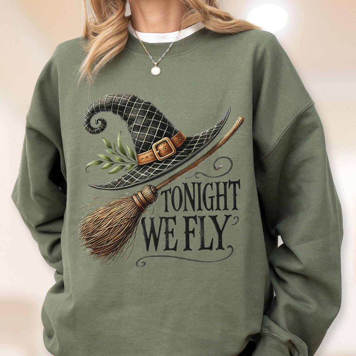 May include: An olive green crewneck sweatshirt featuring a graphic print of a black witch hat with a brown buckle and green leaves, paired with a wooden broomstick. The text "TONIGHT WE FLY" is printed in a dark, stylized serif font below the hat.