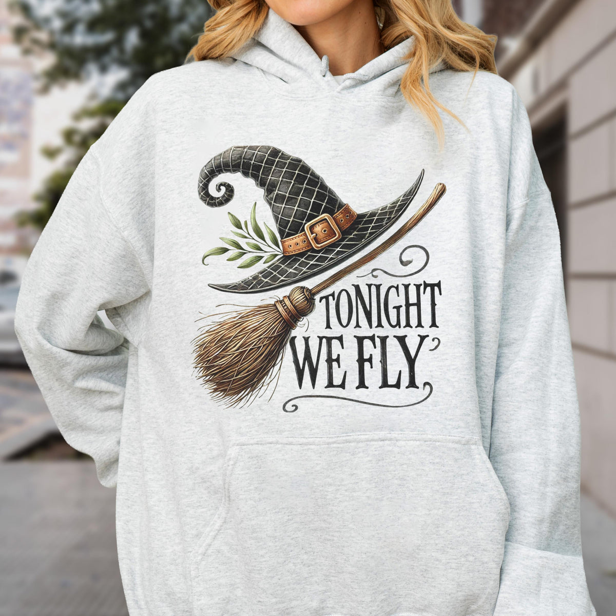 May include: A light gray heathered hoodie featuring a graphic print of a black witch hat with a brown buckle and green leaves, a wooden broomstick, and the black text "TONIGHT WE FLY" written in a stylized serif font on the front center.