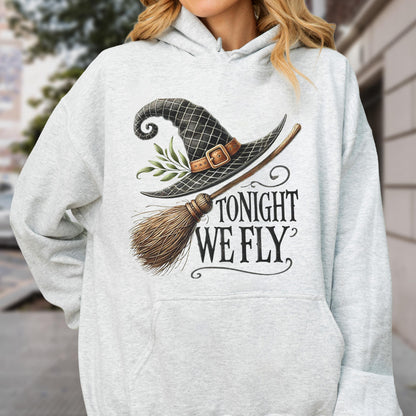 May include: A light gray heathered hoodie featuring a graphic print of a black witch hat with a brown buckle and green leaves, a wooden broomstick, and the black text "TONIGHT WE FLY" written in a stylized serif font on the front center.