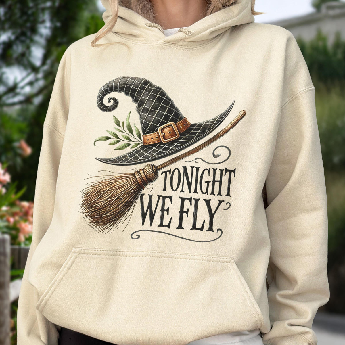 May include: A cream-colored hooded sweatshirt featuring a graphic print of a black witch hat with a brown buckle, a green leafy sprig, and a wooden broomstick. The text "TONIGHT WE FLY" is printed in a dark, stylized serif font below the graphic.