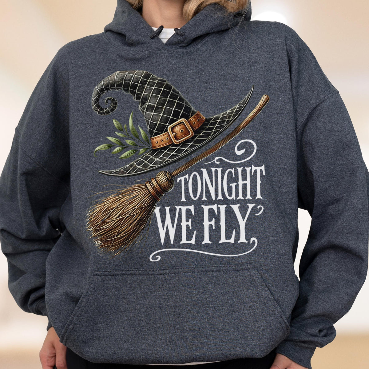 May include: A dark heather gray hooded sweatshirt featuring a graphic print of a black witch hat with a brown buckle, green leaves, and a wooden broomstick. The text TONIGHT WE FLY is printed in white lettering below the hat and broomstick illustration.