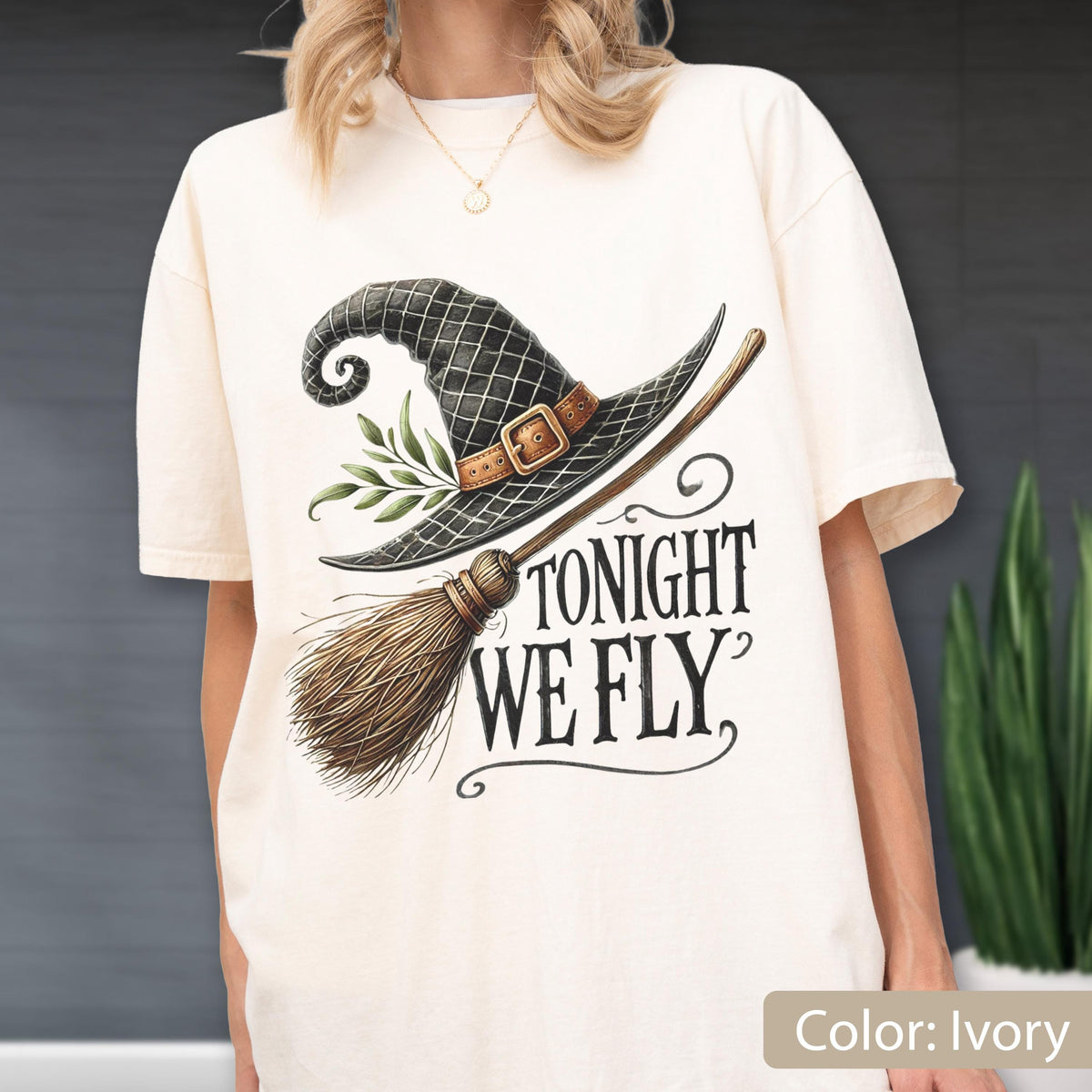 May include: An ivory t-shirt featuring a graphic of a black witch hat with a brown buckle and green leaves, paired with a wooden broomstick. The text TONIGHT WE FLY is printed in black, stylized lettering below the hat and broomstick illustration.