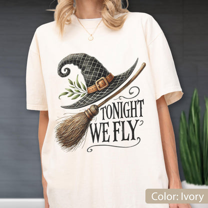 May include: An ivory t-shirt featuring a graphic of a black witch hat with a brown buckle and green leaves, paired with a wooden broomstick. The text TONIGHT WE FLY is printed in black, stylized lettering below the hat and broomstick illustration.