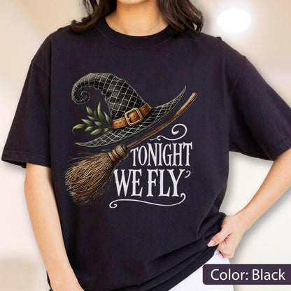 May include: A black t-shirt featuring a graphic print of a black witch hat with a brown buckle and green leaves, paired with a wooden broomstick. The text "TONIGHT WE FLY" is printed in white lettering below the hat and broomstick illustration.