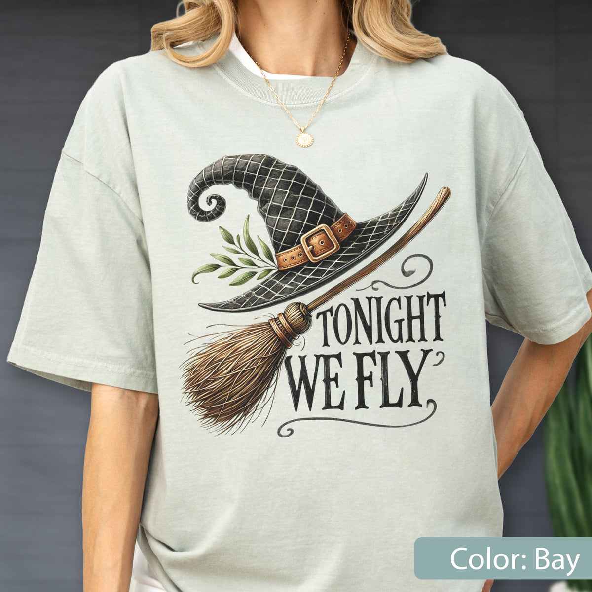 May include: A light bay-colored t-shirt featuring a graphic of a black witch hat with a brown buckle and green leaves, paired with a wooden broomstick. The text TONIGHT WE FLY is printed in a black serif font to the right of the broomstick.