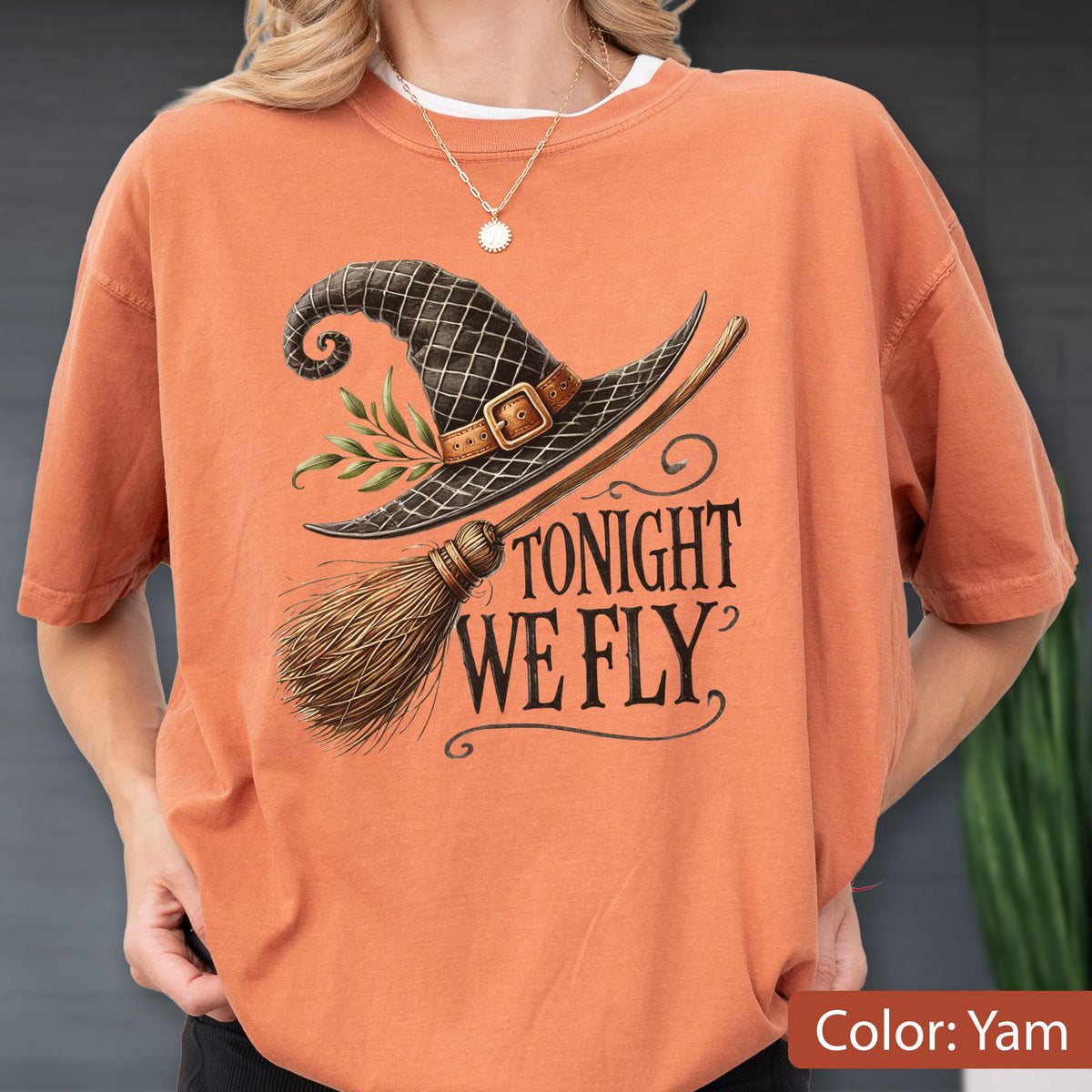 May include: A yam-colored short-sleeve t-shirt featuring a graphic of a black witch hat with a brown buckle and green leaves, paired with a wooden broomstick. The text "TONIGHT WE FLY" is printed in a dark, stylized serif font below the hat.