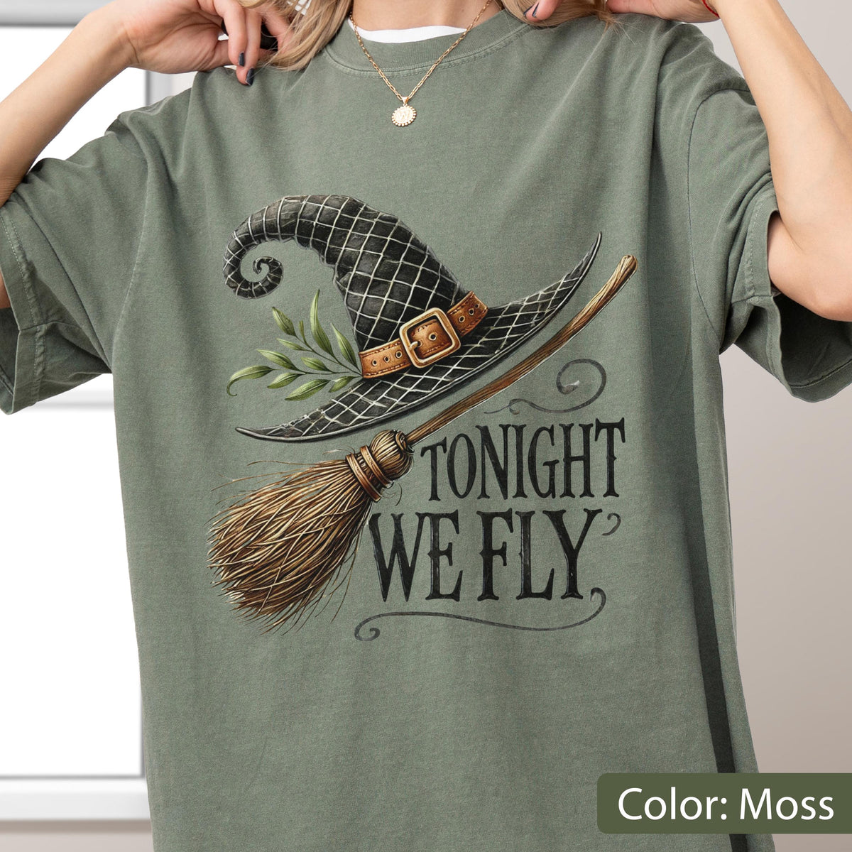 May include: A moss green t-shirt featuring a graphic of a black witch hat with a brown buckle, a sprig of green leaves, and a wooden broomstick. The text "TONIGHT WE FLY" is printed in a dark, stylized serif font to the right of the broom.