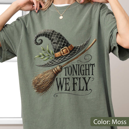 May include: A moss green t-shirt featuring a graphic of a black witch hat with a brown buckle, a sprig of green leaves, and a wooden broomstick. The text "TONIGHT WE FLY" is printed in a dark, stylized serif font to the right of the broom.