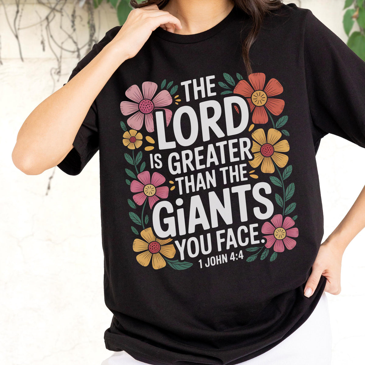 May include: A black short-sleeved t-shirt featuring a graphic print with the text "THE LORD IS GREATER THAN THE GIANTS YOU FACE. 1 JOHN 4:4" in white, surrounded by a colorful floral border with pink, yellow, and orange flowers and green leaves.