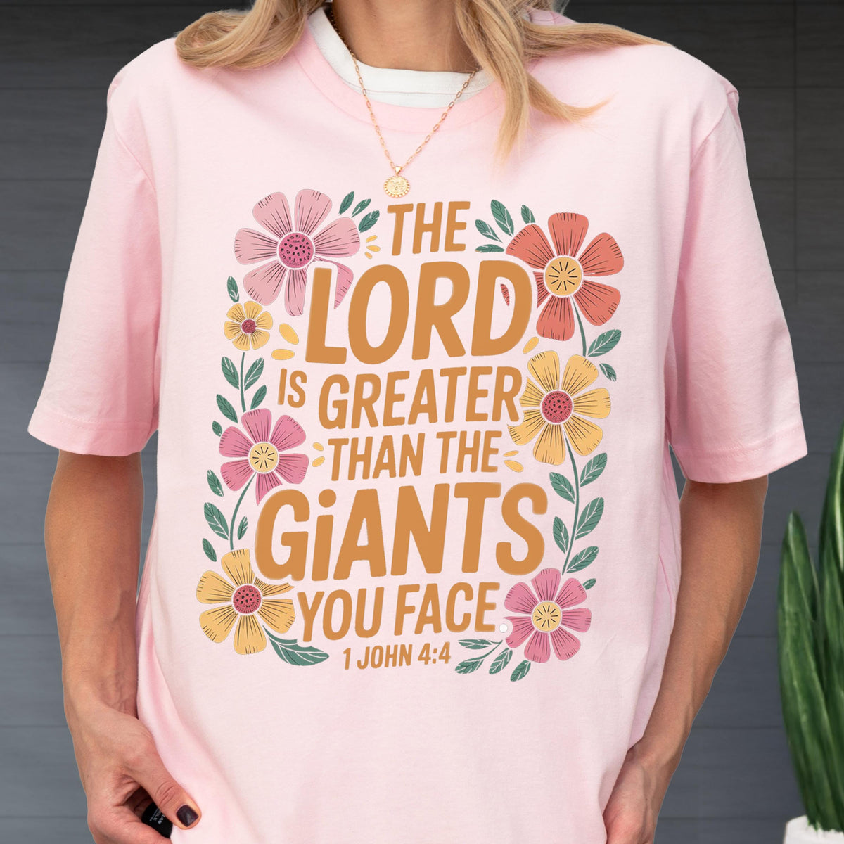 May include: A light pink short-sleeved t-shirt featuring the text THE LORD IS GREATER THAN THE GIANTS YOU FACE 1 JOHN 4:4 in brown lettering, surrounded by a decorative border of colorful pink, orange, and yellow flowers with green leaves.