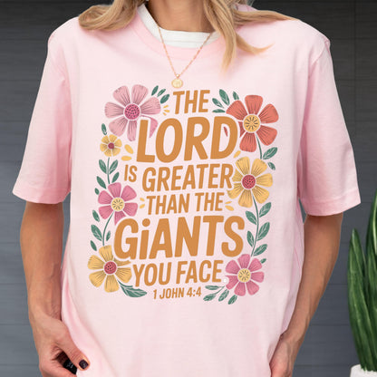 May include: A light pink short-sleeved t-shirt featuring the text THE LORD IS GREATER THAN THE GIANTS YOU FACE 1 JOHN 4:4 in brown lettering, surrounded by a decorative border of colorful pink, orange, and yellow flowers with green leaves.