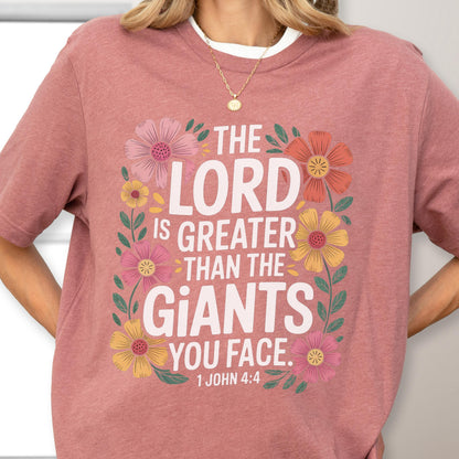 May include: A dusty rose short-sleeved t-shirt featuring a floral border design surrounding the white text: THE LORD IS GREATER THAN THE GIANTS YOU FACE. 1 JOHN 4:4. The flowers are in shades of pink, yellow, and orange with green leaves.