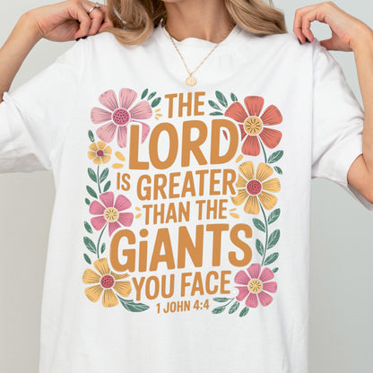 May include: A white short-sleeved t-shirt featuring a graphic print with the text "THE LORD IS GREATER THAN THE GIANTS YOU FACE 1 JOHN 4:4" in brown lettering, surrounded by a decorative border of pink, orange, and yellow flowers with green leaves.