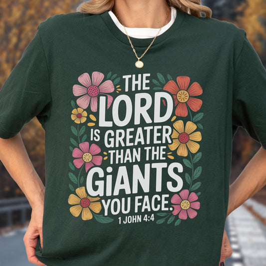 May include: A dark green short-sleeve t-shirt featuring a floral graphic design with the white text "THE LORD IS GREATER THAN THE GIANTS YOU FACE. 1 JOHN 4:4" surrounded by pink, orange, and yellow flowers with green leaves on the front of the garment.