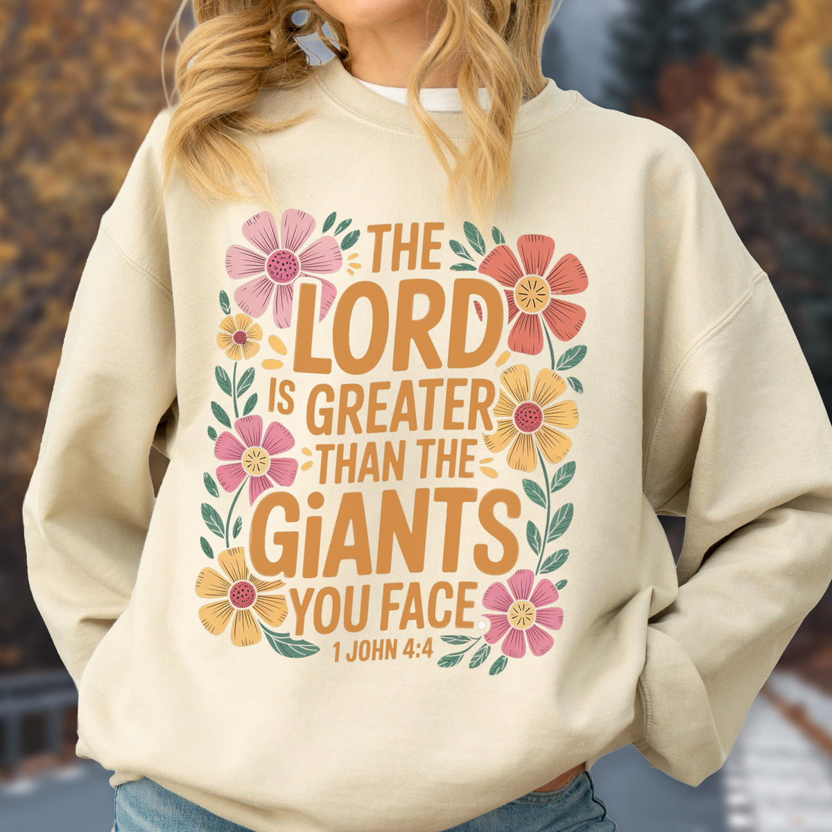 May include: Beige sweatshirt with the text "THE LORD IS GREATER THAN THE GIANTS YOU FACE" in orange and white, surrounded by pink, orange, and yellow floral accents. The scripture reference "1 JOHN 4:4" is at the bottom.