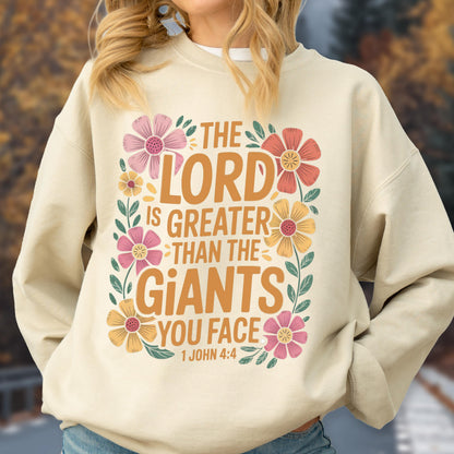 May include: Beige sweatshirt with the text "THE LORD IS GREATER THAN THE GIANTS YOU FACE" in orange and white, surrounded by pink, orange, and yellow floral accents. The scripture reference "1 JOHN 4:4" is at the bottom.