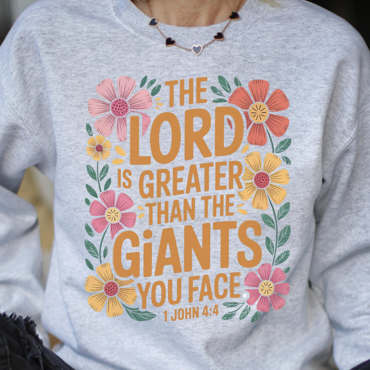 May include: A light gray sweatshirt with the text "THE LORD IS GREATER THAN THE GIANTS YOU FACE" in orange, surrounded by colorful floral designs. The text "1 JOHN 4:4" is at the bottom.