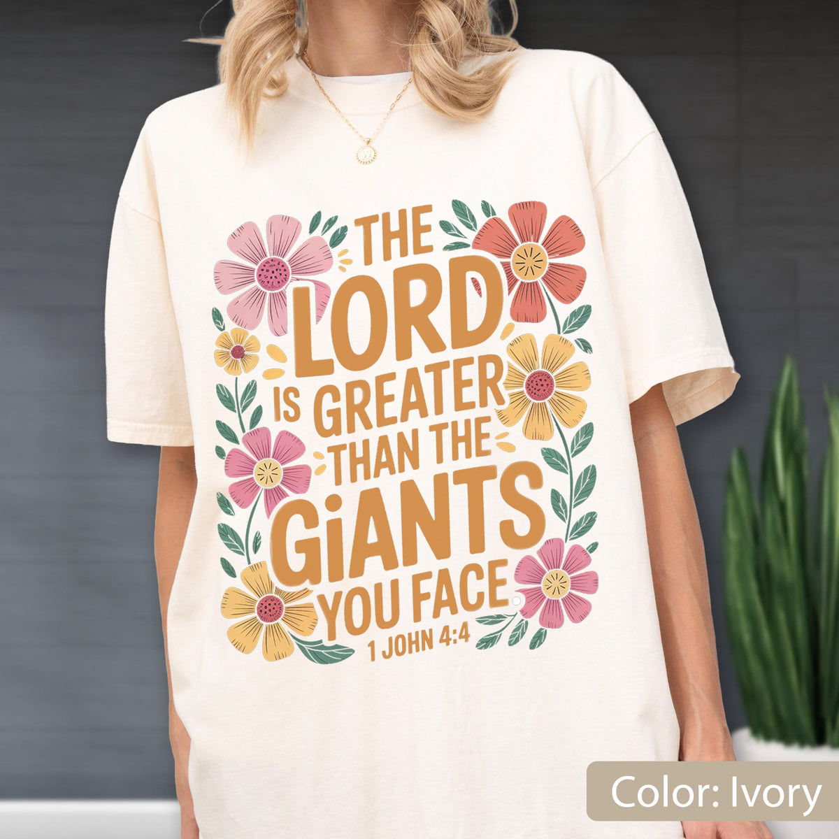May include: Ivory-colored t-shirt with the text "THE LORD IS GREATER THAN THE GIANTS YOU FACE 1 JOHN 4:4" in a floral design. The design includes pink, orange, and yellow flowers with green leaves.