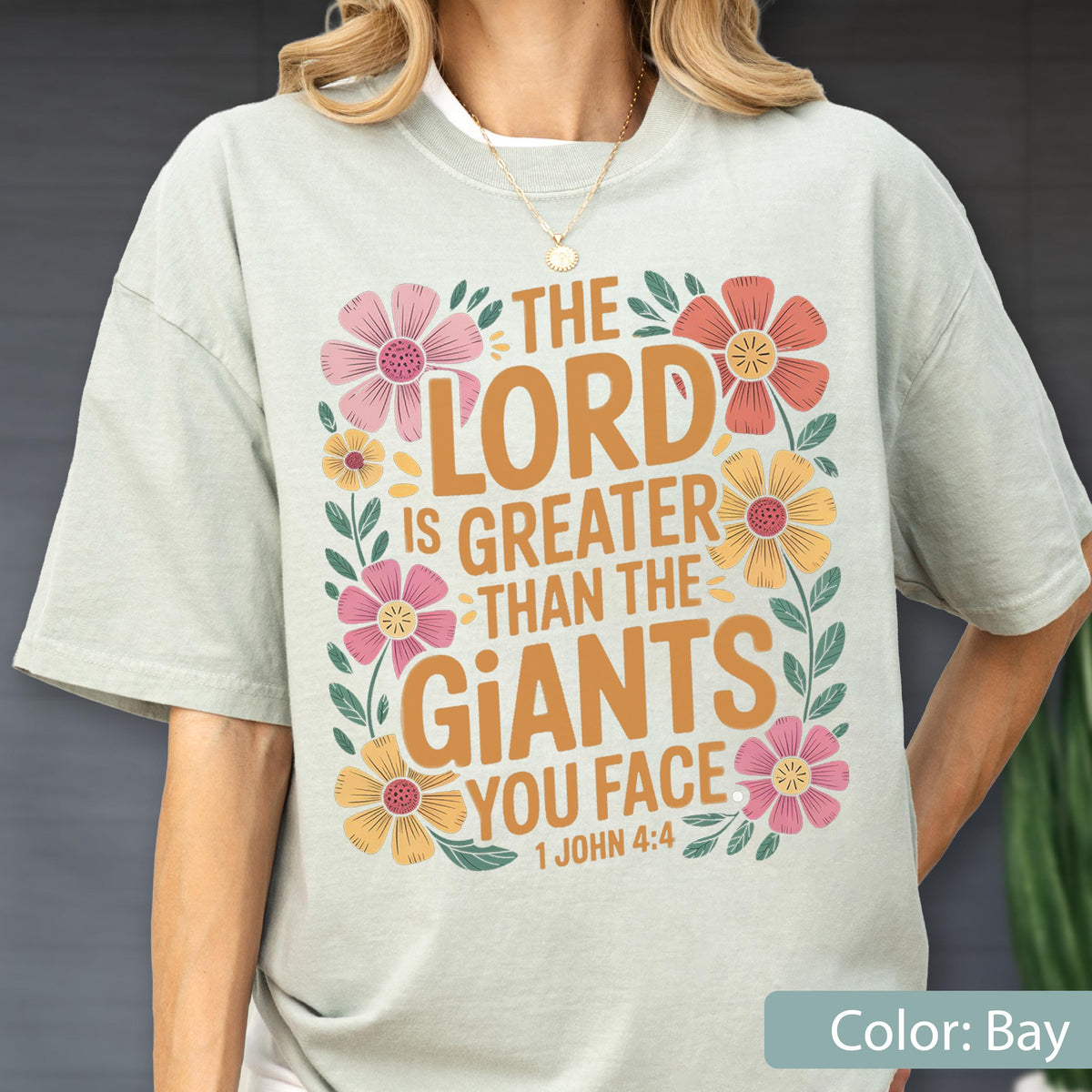 May include: A light green t-shirt with the text "THE LORD IS GREATER THAN THE GIANTS YOU FACE 1 JOHN 4:4" in orange. The text is surrounded by colorful floral illustrations in pink, yellow, and orange.