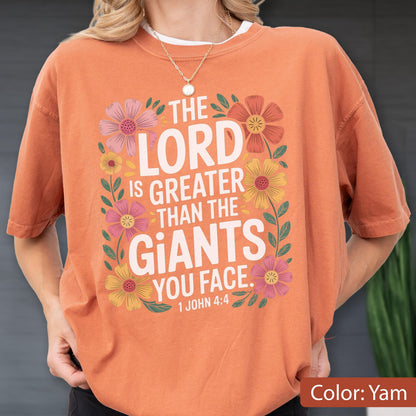 May include: Orange t-shirt with the text "THE LORD IS GREATER THAN THE GIANTS YOU FACE. 1 JOHN 4:4" surrounded by colorful flowers. The shirt is a solid color, and the text is in a bold, readable font.