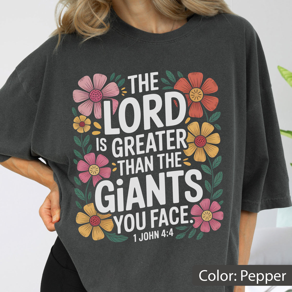 May include: Pepper-colored t-shirt with the text "THE LORD IS GREATER THAN THE GIANTS YOU FACE. 1 JOHN 4:4" in white. The text is surrounded by colorful floral illustrations in pink, orange, and yellow.