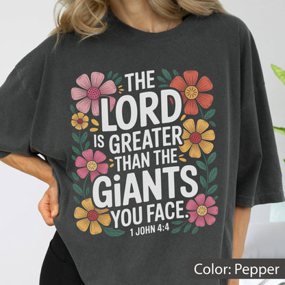 May include: Pepper-colored t-shirt with the text "THE LORD IS GREATER THAN THE GIANTS YOU FACE. 1 JOHN 4:4" in white. The text is surrounded by colorful floral illustrations in pink, orange, and yellow.