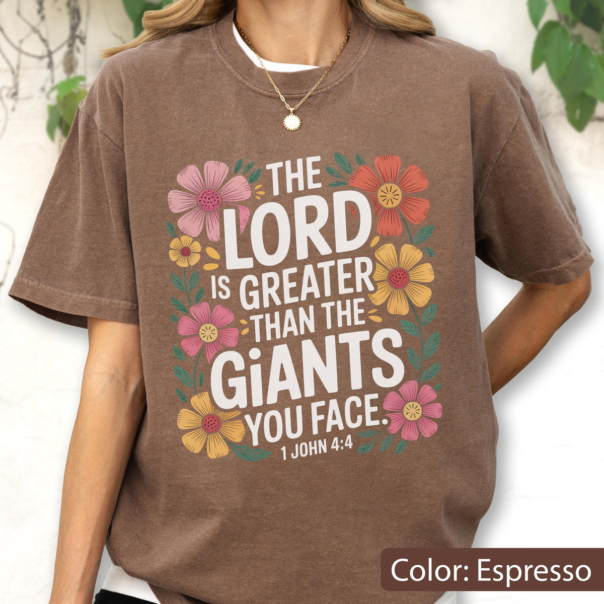 May include: Espresso-colored t-shirt with the text "THE LORD IS GREATER THAN THE GIANTS YOU FACE. 1 JOHN 4:4" surrounded by colorful floral graphics. The shirt has short sleeves and a crew neck.