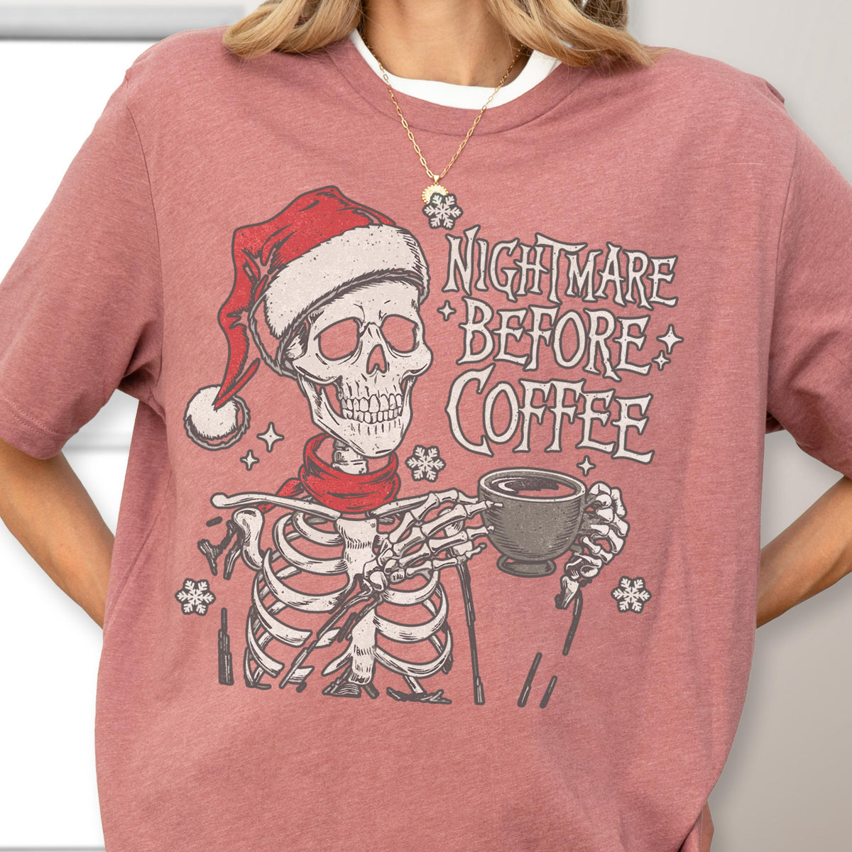 May include: A mauve-colored t-shirt featuring a skeleton wearing a Santa hat and a red scarf, holding a coffee cup. The text "NIGHTMARE BEFORE COFFEE" is printed above the skeleton. The design includes snowflake and star accents.