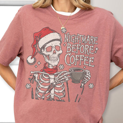 May include: A mauve-colored t-shirt featuring a skeleton wearing a Santa hat and a red scarf, holding a coffee cup. The text "NIGHTMARE BEFORE COFFEE" is printed above the skeleton. The design includes snowflake and star accents.