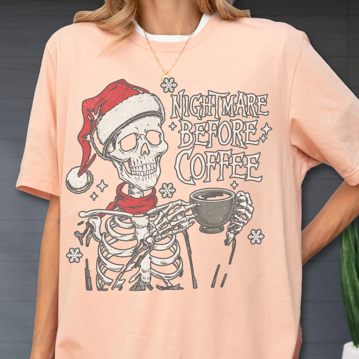 May include: Peach-colored t-shirt featuring a skeleton wearing a Santa hat and red scarf, holding a coffee cup. The text "Nightmare Before Coffee" is printed above the skeleton. Snowflake graphics are also present.