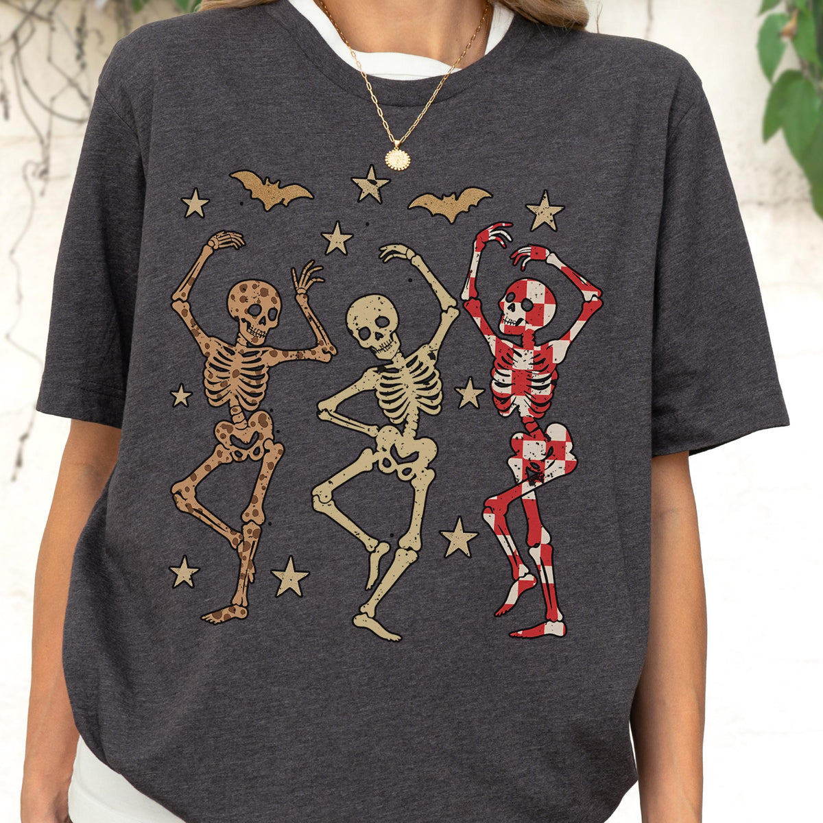 May include: Dark gray t-shirt featuring a vintage-style graphic of three dancing skeletons. The skeletons are in various colors, with bats and stars scattered around them. A fun and spooky design for Halloween.