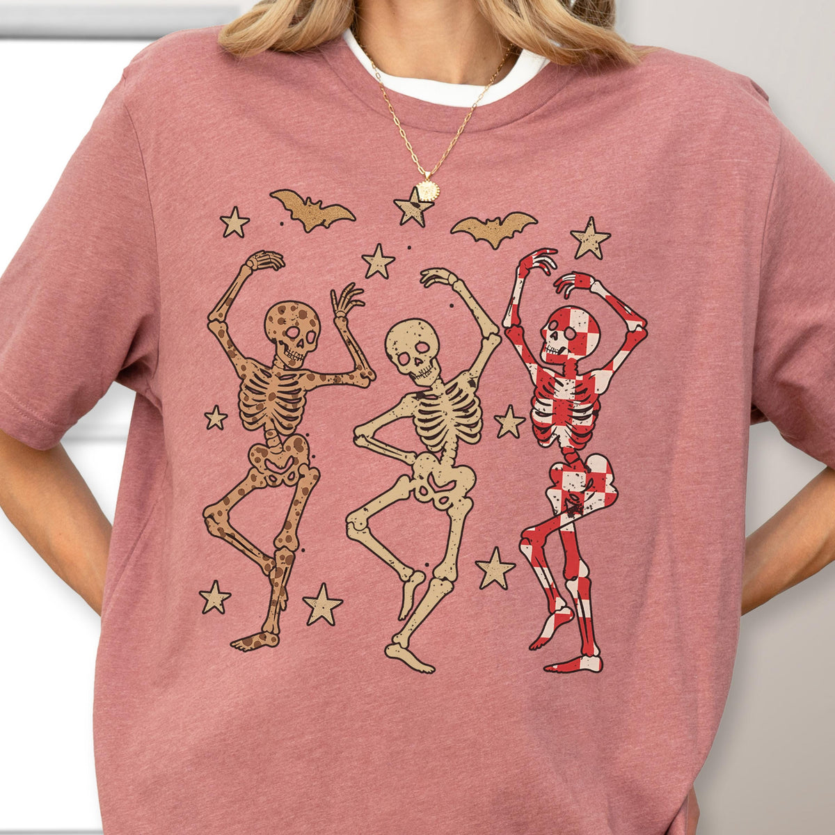 May include: A mauve-colored t-shirt featuring a graphic of three dancing skeletons. The skeletons are in various colors, including brown, beige, and red, with star and bat accents. The design is a playful, Halloween-themed illustration.
