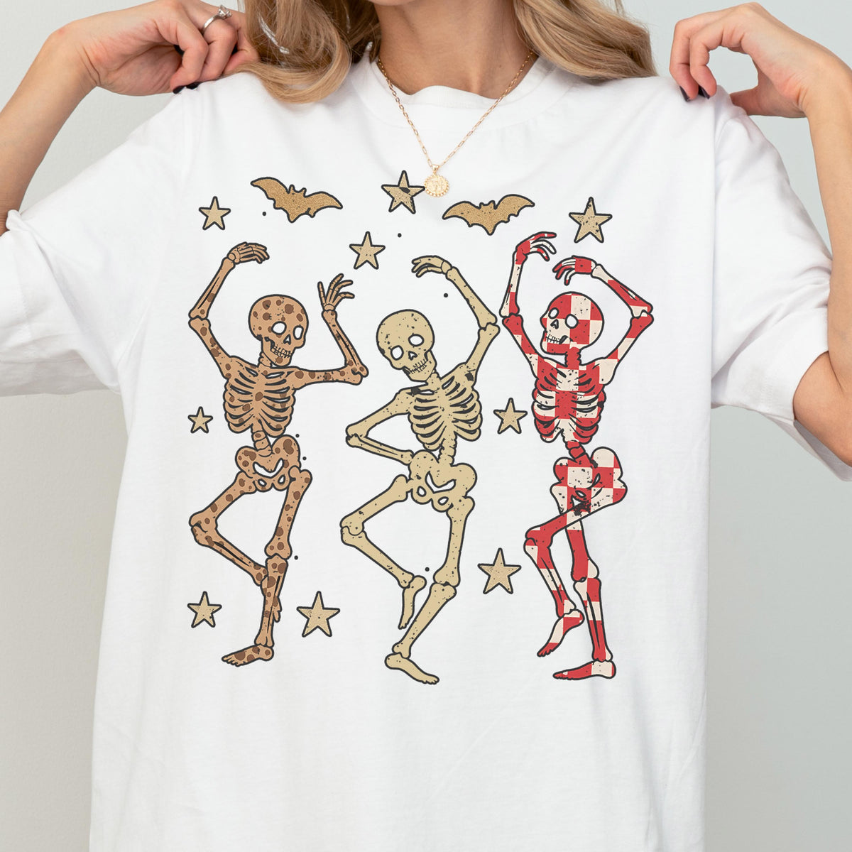 May include: White t-shirt featuring a playful Halloween design. The graphic includes three dancing skeletons in various colors, surrounded by stars and bats. The skeletons have different patterns, including brown, beige, and red and white checkered designs.