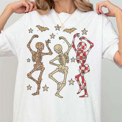 May include: White t-shirt featuring a playful Halloween design. The graphic includes three dancing skeletons in various colors, surrounded by stars and bats. The skeletons have different patterns, including brown, beige, and red and white checkered designs.