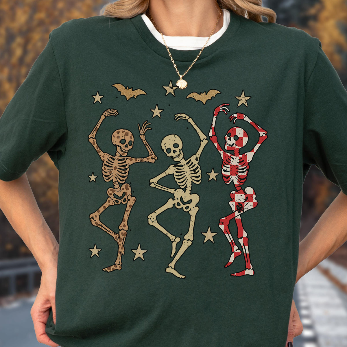 May include: Dark green t-shirt featuring a graphic of three dancing skeletons. The skeletons are in various colors, including tan and red checkered patterns. Bats and stars are also included in the design. The shirt has a round neckline and short sleeves.