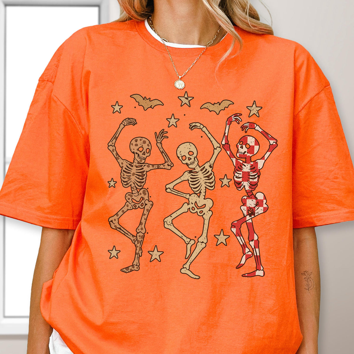 May include: Orange t-shirt featuring a graphic of three dancing skeletons. The skeletons are in various poses, with star and bat illustrations above them. The skeleton on the right has a red and white checkered pattern. A round neck shirt.